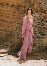 Vestido largo de gasa color rosa, con escote en V, mangas largas abullonadas y volantes en el frente y en la falda. Incluye forro interior y cierre de lazo en la parte delantera.