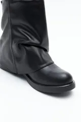 Bota de caña baja color negro, confeccionada en poliuretano con acabado liso y estructura flexible que genera un efecto fruncido. Presenta puntera redondeada, base baja y un cierre metálico en la parte trasera de la caña.