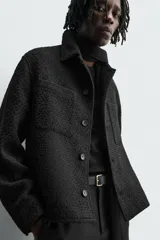 Sobrecamisa de corte relajado con textura de tejido estructurado en color gris y negro. Presenta cuello clásico, cierre frontal con botones, mangas largas con puños abotonados y bolsillos de plastrón en el pecho.