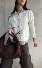 Cardigan de punto trenzado color natural, con cuello redondo, cierre frontal de botones negros y mangas largas con puños acanalados.