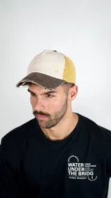 Gorra tipo trucker con visera desgastada, combinando paneles en color beige, marrón y amarillo mostaza. Presenta un pequeño bordado en el panel frontal.