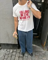 Remera blanca de manga corta, con cuello redondo y diseño estampado en el centro con la frase "Killing in the name of" en color rojo.