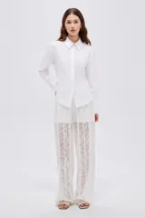 Camisa blanca de algodón, holgada, de manga larga con puños anchos, cuello camisero y cierre frontal oculto con un botón dorado visible en el cuello.