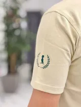 Remera color beige con cuello redondo y mangas cortas. Estampado frontal con el texto "Duxonstudios" en tipografía cursiva, acompañado de un logotipo circular con hojas de laurel y una figura central en tono verde oscuro.
