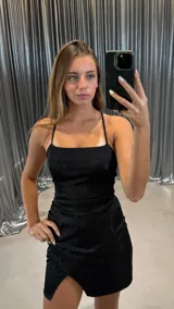 Vestido corto negro con brillo, corte ajustado y escote cuadrado con tirantes finos cruzados en la espalda. La falda presenta un cruce frontal con frunce lateral.