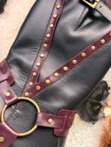Bota alta de cuero negro con taco cuadrado de efecto madera, puntera redondeada y detalles de tiras en tono bordó con tachas metálicas doradas. Cuenta con una argolla metálica en el empeine y una hebilla de ajuste en la parte superior de la caña.