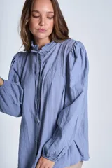 Camisa de color azul con cuello alto decorado con volados y lazo de ajuste. Presenta tablas decorativas en los hombros, cierre frontal con botones de nácar y mangas largas con puños elásticos que generan un efecto abullonado.