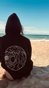 Buzo canguro negro con capucha y cordones ajustables. Estampado en la espalda con un diseño circular de una ola, un sol y una palmera.