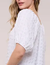 Blusa blanca de tela texturada y silueta fluida, con escote en U y frunces. Cuenta con mangas cortas abullonadas con elástico en los puños y ruedo redondeado.