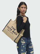 Bolso tipo tote de Marc Jacobs, modelo The Jacquard Large Tote Bag, confeccionado en tejido jacquard color tostado. Presenta el nombre de la marca y el modelo bordados en el frente en color blanco, doble asa superior y detalles de cintas laterales con inscripciones.