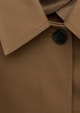 Chaqueta larga de algodón color marrón tostado, estilo gabardina, con cuello camisero, cierre frontal oculto con botones, mangas largas con presillas en los puños y cinturón ajustable con hebilla oscura.