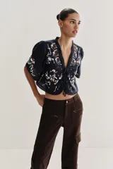 Top corto de color negro con escote en pico y mangas cortas anchas. Presenta bordados coloridos en estilo geométrico y floral, detalle de entredoses y cierre frontal mediante lazos con abalorios.