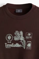 Buzo marrón de corte holgado con cuello redondo, confeccionado en mezcla de algodón. Presenta un estampado gráfico frontal con motivos botánicos y la inscripción "Taxodiaceae". Cuenta con puños y bajo acanalados.