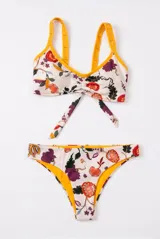 Conjunto de bikini de dos piezas con estampado floral en tonos rojo, violeta y verde sobre fondo claro, con detalles en color amarillo en los bordes y tirantes. El corpiño es de estilo deportivo con tiras para anudar al frente, y la bombacha es de corte clásico.