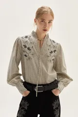 Blusa de algodón con estampado de rayas verticales en tonos crudo y negro. Presenta cuello mao con lazo para atar, bordados florales en negro y beige en la zona del pecho y hombros, y mangas largas con puños fruncidos elásticos.