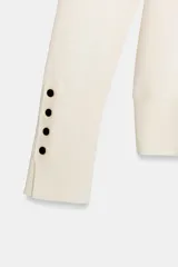 Jersey de punto liso en color beige, con cuello alto y manga larga con detalle de botones metálicos en los puños.