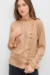 Remera de manga larga color beige, con cuello alto y cierre frontal asimétrico con botones. Presenta detalles de volados en el cuello, a lo largo de la abotonadura y en los puños. Confeccionada en un tejido liviano con caída fluida.