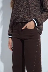 Pantalón de denim corte wide leg color marrón, con ruedo desflecado y detalle de tachas metálicas a lo largo de las costuras laterales.