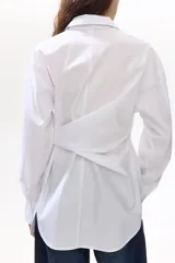 Camisa de algodón blanca, de manga larga con puños abotonados, cuello camisero y cierre frontal de botones. Presenta un diseño con tiras incorporadas en el lateral para anudar, creando un efecto cruzado y drapeado en el frente.