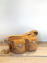 Bolso de hombro confeccionado en gamuza color marrón, con diseño de dos bolsillos frontales con solapa y cierres metálicos tipo broche. Presenta costuras a la vista y una correa ancha con detalles de remaches metálicos.