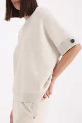 Sweater de punto color beige, con cuello redondo, mangas cortas con dobladillo y detalle de botón lateral, y terminaciones en lurex.