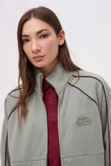 Campera de felpa con friza, color verde oliva, con cierre metálico frontal, cuello alto y detalles de vivos en color bordó. Presenta un bordado en el pecho y corte relaxed.