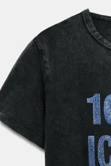 Remera negra de efecto lavado con corte slim fit, cuello redondo y manga corta. Cuenta con un estampado frontal con la frase "100% ICONIC" hecha con aplicaciones de brillos plateados.