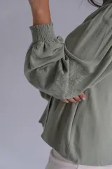 Blusa de color verde agua, con cuello en V, cierre frontal con botones y mangas largas abullonadas con puños elásticos. Presenta detalles bordados sutiles en la parte frontal y en las mangas.