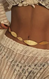Cinto de cadena dorado, estilo body chain, compuesto por eslabones metálicos con forma de hoja alargada y textura martillada. Se ajusta a la cintura.