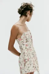Vestido corto strapless de corte corsetero, con estampado floral en tonos rosados y verdes sobre fondo blanco. Presenta detalles de tiras cruzadas ajustables en el delantero con lazada y varillas interiores.