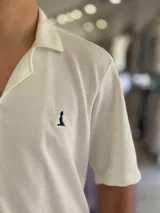 Camisa de manga corta color blanco marfil, confeccionada en tejido texturizado con cuello camisero, cierre frontal de botones y un pequeño bordado oscuro en el pecho.