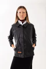 Campera de cuero negro con cuello clásico, cierre frontal oculto y bolsillos laterales. Presenta un parche de cuero con el logo de la marca en la parte inferior.