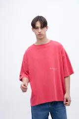 Remera de algodón teñida en prenda, color coral/rosado, con calce boxy y estampado en la espalda con texto en tipografía cursiva y líneas de texto más pequeñas.