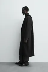 Abrigo largo de corte relajado en color negro, confeccionado en tejido con viscosa. Presenta un diseño de botonadura cruzada, cuello con solapas de pico, manga larga con puños abotonados y bolsillos laterales de vivo. Incluye una abertura en el centro de la espalda y un bolsillo interior.