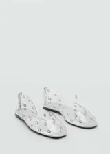 Zapato plano transparente con detalle de tachas metálicas, punta fina y tira ajustable con hebilla en el empeine.