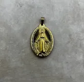 Dije ovalado de metal dorado con la imagen en relieve de la Virgen Milagrosa y una inscripción perimetral. Posee un diseño clásico y elegante con un tamaño aproximado de 4,5 cm.
