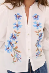 Camisa blanca de manga larga abullonada, con delicados bordados florales en tonos azules y verdes a lo largo de la botonadura frontal y en las mangas. Presenta cuello mao con reborde en contraste y cierre frontal con botones.