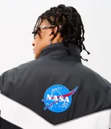 Campera deportiva de poliéster con diseño de bloques de color en negro y blanco. Presenta cuello alto, cierre frontal con cremallera y parches de la NASA y la bandera de Estados Unidos en el pecho.
