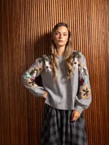 Buzo de cuello redondo en color gris, con mangas largas y diseño holgado. Presenta un bordado floral multicolor con detalles de cuentas en la parte frontal y en las mangas.