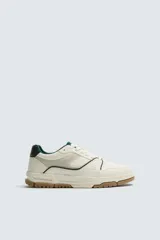 Zapatilla deportiva de estilo retro con diseño de paneles en tonos crema y blanco, detalles en verde oscuro y suela de goma color caramelo.