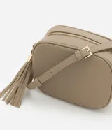 Cartera bandolera pequeña color beige, confeccionada en poliuretano con textura granulada. Presenta cierre superior con cremallera, correa larga ajustable y detalle de borla decorativa en el lateral con herrajes dorados.
