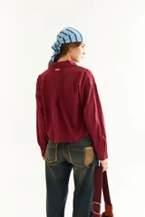 Camisa de corte boxy y calce holgado, color bordó, con cuello clásico, cierre frontal de botones y detalle de tablas con recortes diagonales en el pecho.