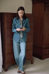 Camisa de denim azul de manga larga con cuello solapa y cierre frontal de botones. Presenta bajo redondeado.