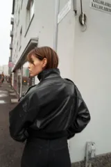 Cazadora de estilo bomber en efecto piel color negro, con cuello subido, manga larga y bajo elástico. Presenta trabillas en los hombros, bolsillos frontales con solapa y cierre de cremallera con botones a presión ocultos.