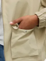 Campera tipo parka corta de color beige, con capucha ajustable con cordón, cierre frontal y grandes bolsillos frontales con solapa. Presenta puños elásticos fruncidos.