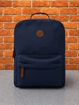 Mochila de lona color azul marino con compartimento principal y bolsillo frontal con cierre. Presenta detalles en cuero marrón, incluyendo un parche circular con logo en la parte superior y tiradores de cierre. Cuenta con asa superior de mano.