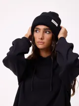 Gorro tipo beanie de tejido acanalado color negro, con vuelta y etiqueta rectangular con logo de la marca en el frente.