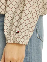 Campera deportiva Tommy Hilfiger color beige con estampado de logos en marrón. Tiene cuello alto, cierre de cremallera, dos bolsillos laterales con botones, puños elásticos y logo bordado en el puño.