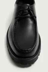 Zapato de cordones de estilo derby, color negro, con parte superior lisa, detalle de bordón en el empeine, horma redonda y suela track dentada.