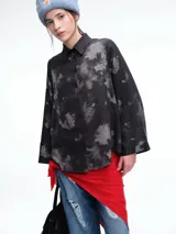 Camisa oversize de lyocell con estampado orgánico efecto lavado. Presenta cuello clásico, cierre frontal con botones, mangas amplias con puños anchos y un diseño de corte relajado con ruedo asimétrico, siendo más largo en la parte posterior.
